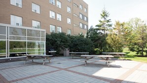 An outdoor patio with picnic tables, a brick building with large windows, and a grassy area with trees. - Residence & Conference Centre - Windsor (Windsor)