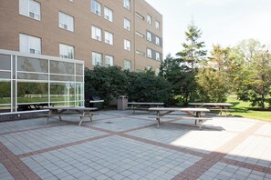 Un patio al aire libre con mesas de picnic, un edificio de ladrillo con ventanas grandes y una zona verde con árboles.