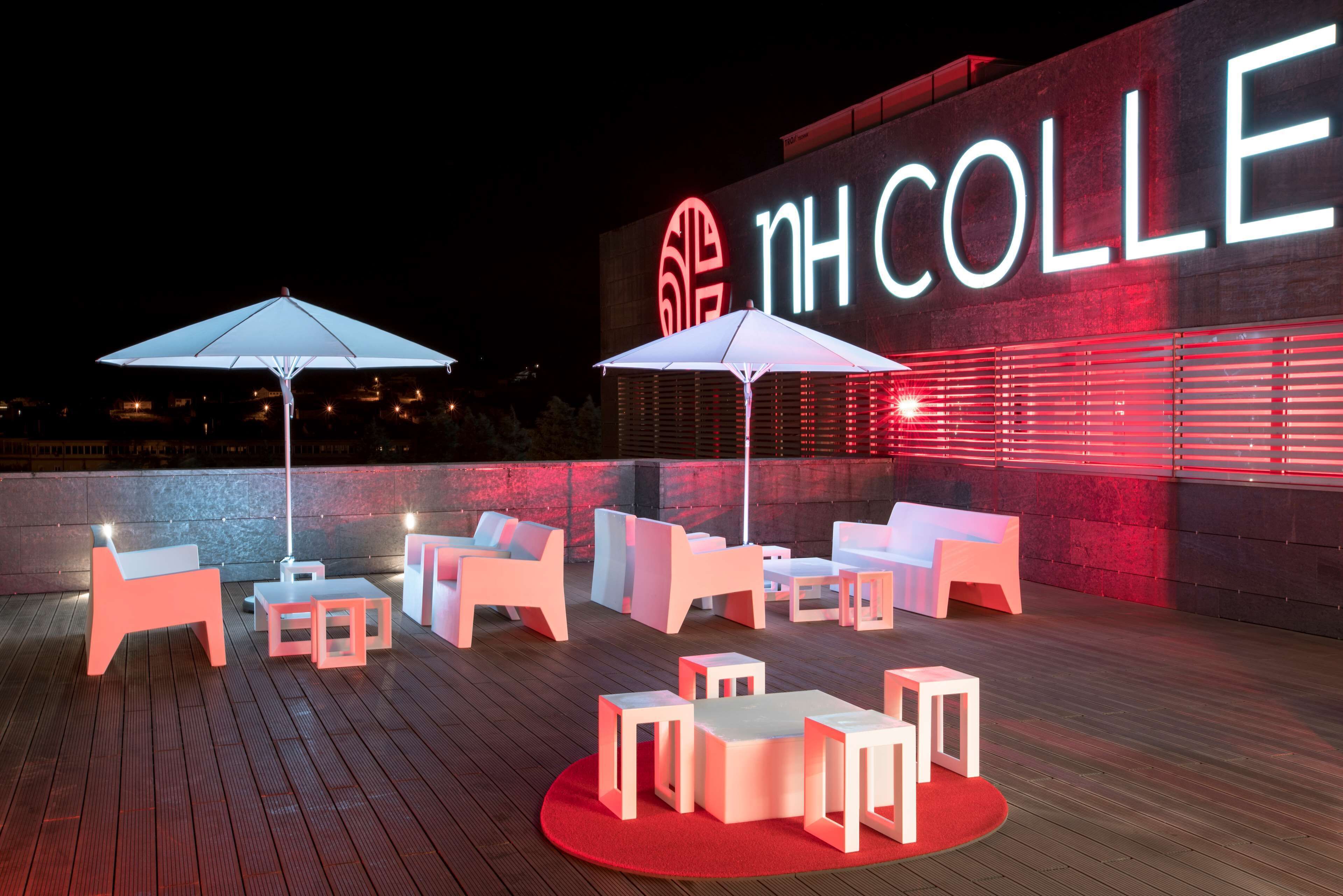 Un área de estar al aire libre con mobiliario moderno y sombrillas, una pared iluminada en rojo con el texto 'NH COLLE', y un cielo nocturno despejado.