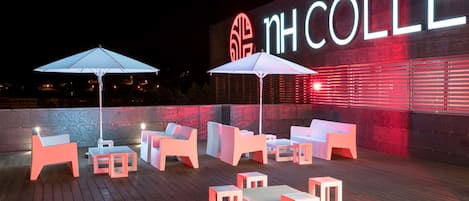 Un espace extérieur aménagé avec des meubles modernes et des parasols, un mur illuminé en rouge sur lequel on peut lire le texte « NH COLLE », et un ciel nocturne dégagé.