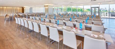 A conference room with rows of tables and chairs, large windows with a city view, and a wooden floor.