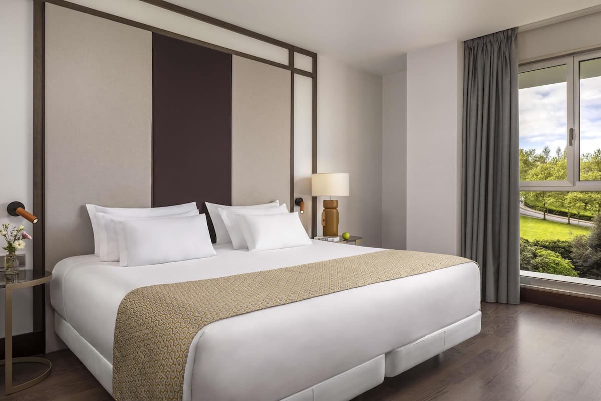 Hotelkamer met een groot bed, een nachtkastje met een lamp en uitzicht op groen door het raam.