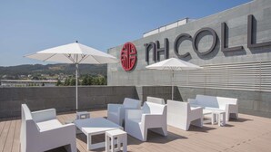A rooftop terrace with white furniture and umbrellas, a large sign with "NH COLL" on a building, and a view of a town in the background.