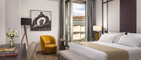A hotel room with a large bed, a small desk with a chair, a bench, a side table with a lamp, a vase with flowers, and a framed picture on the wall.