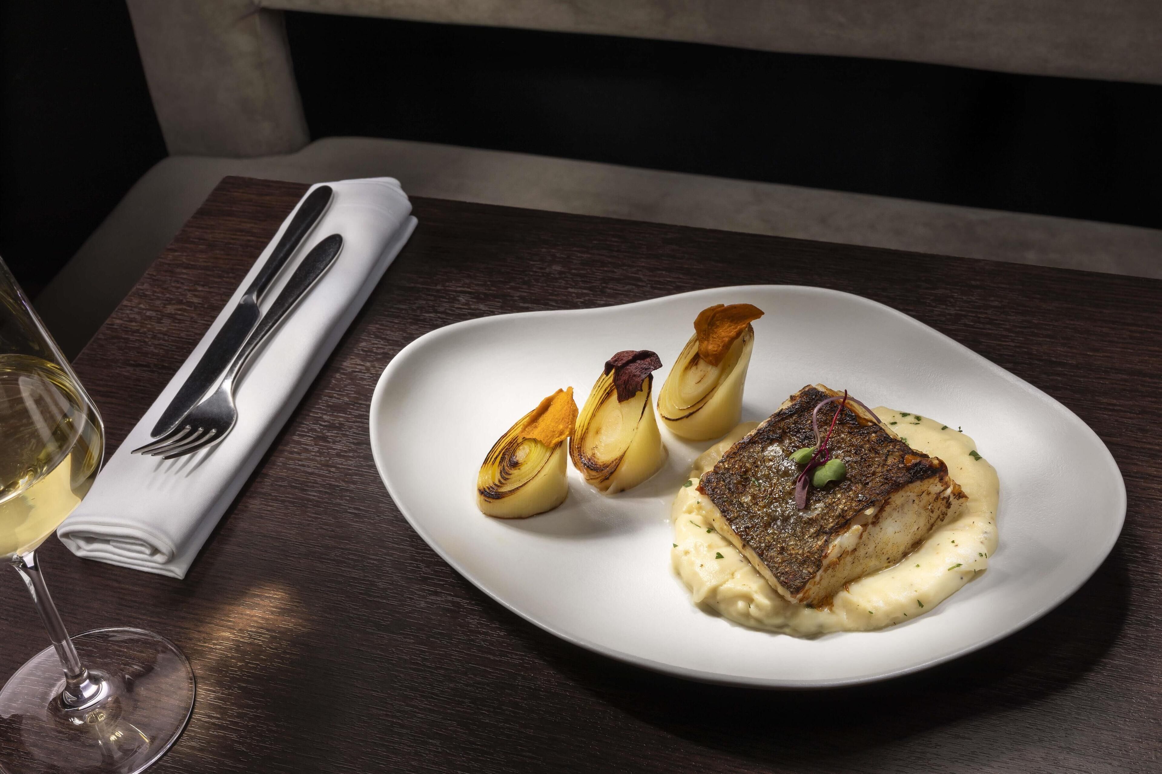 Une assiette blanche contenant un morceau de poisson, des pommes de terre rôties et une sauce crémeuse, le tout posé sur une table en bois sombre.