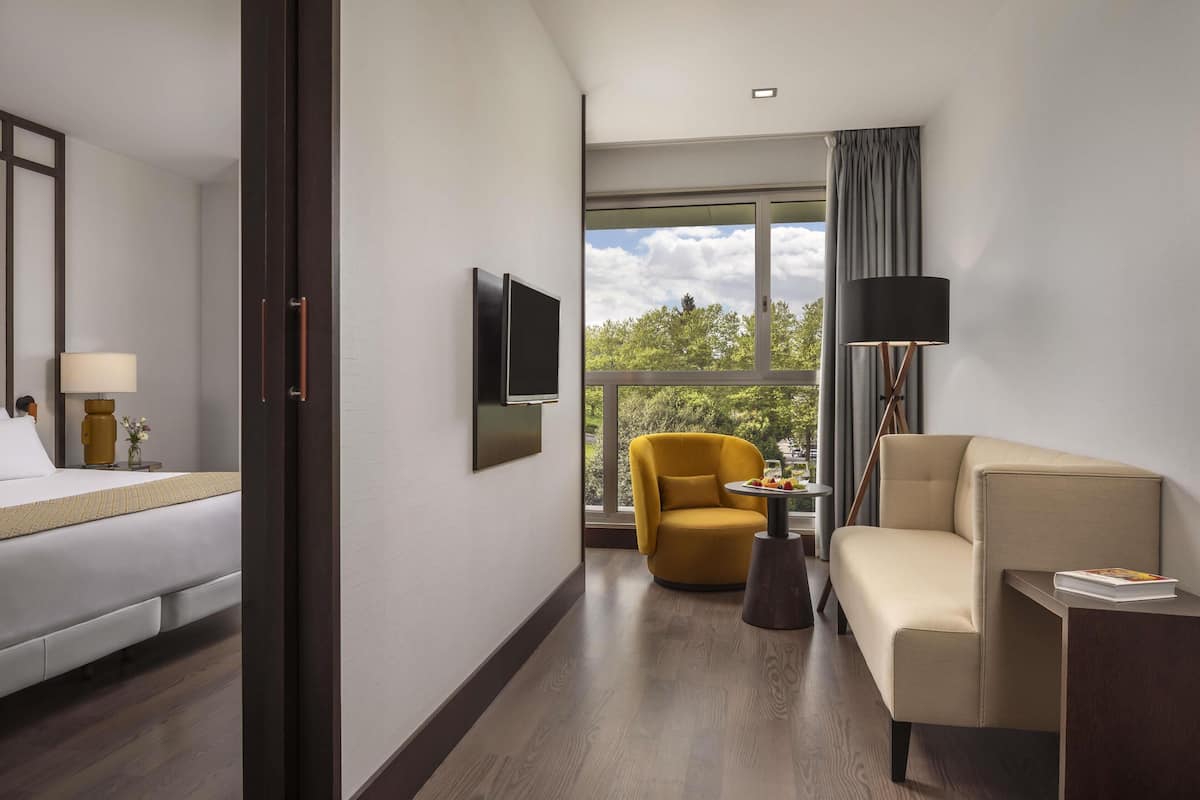 Hotelkamer met een bed, een slaapbank, een gele fauteuil, een aan de muur bevestigde televisie en een groot raam met uitzicht op bomen.