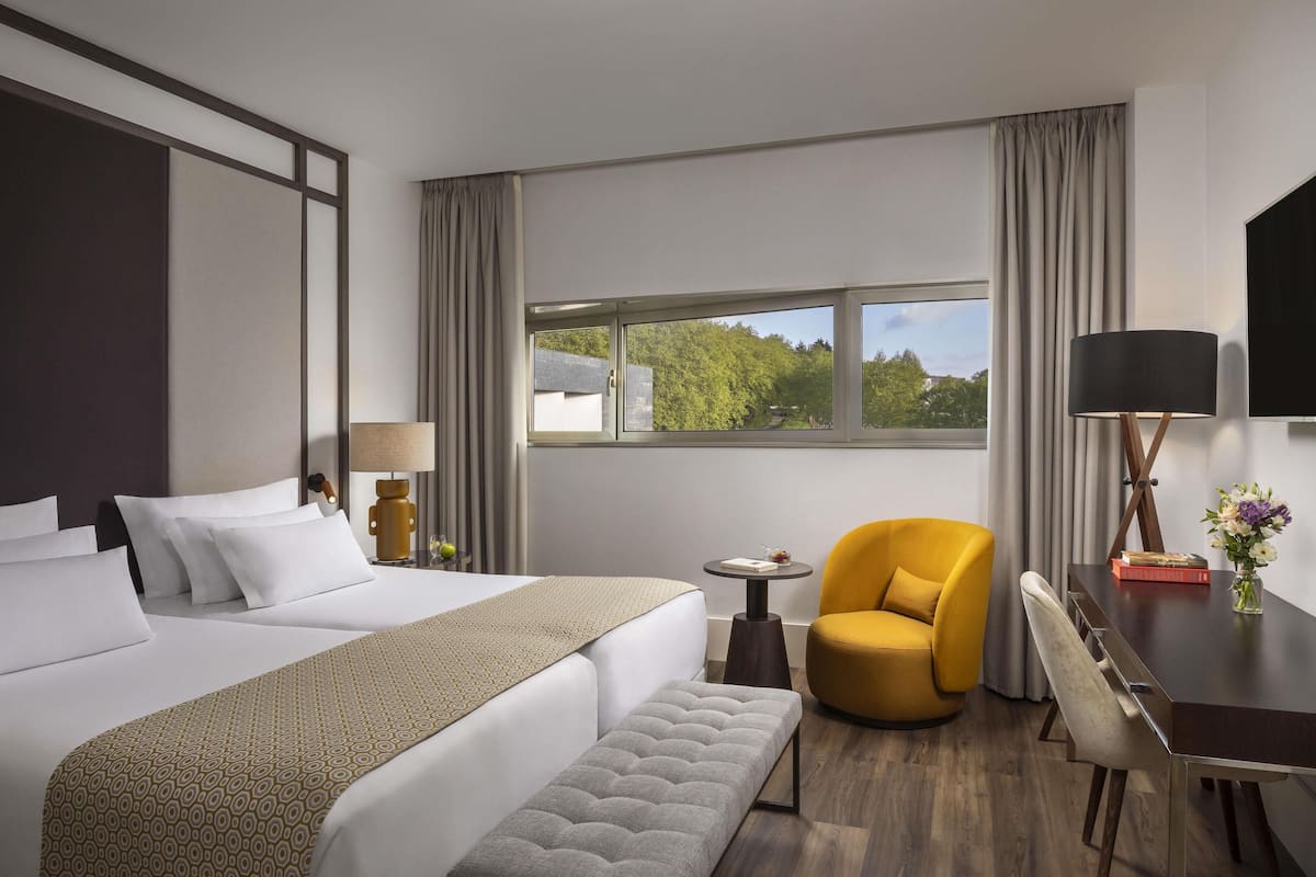Hotelkamer met een groot bed, een bankje, een kleine tafel, een gele fauteuil, een bureau met een lamp, en uitzicht op bomen door het raam.
