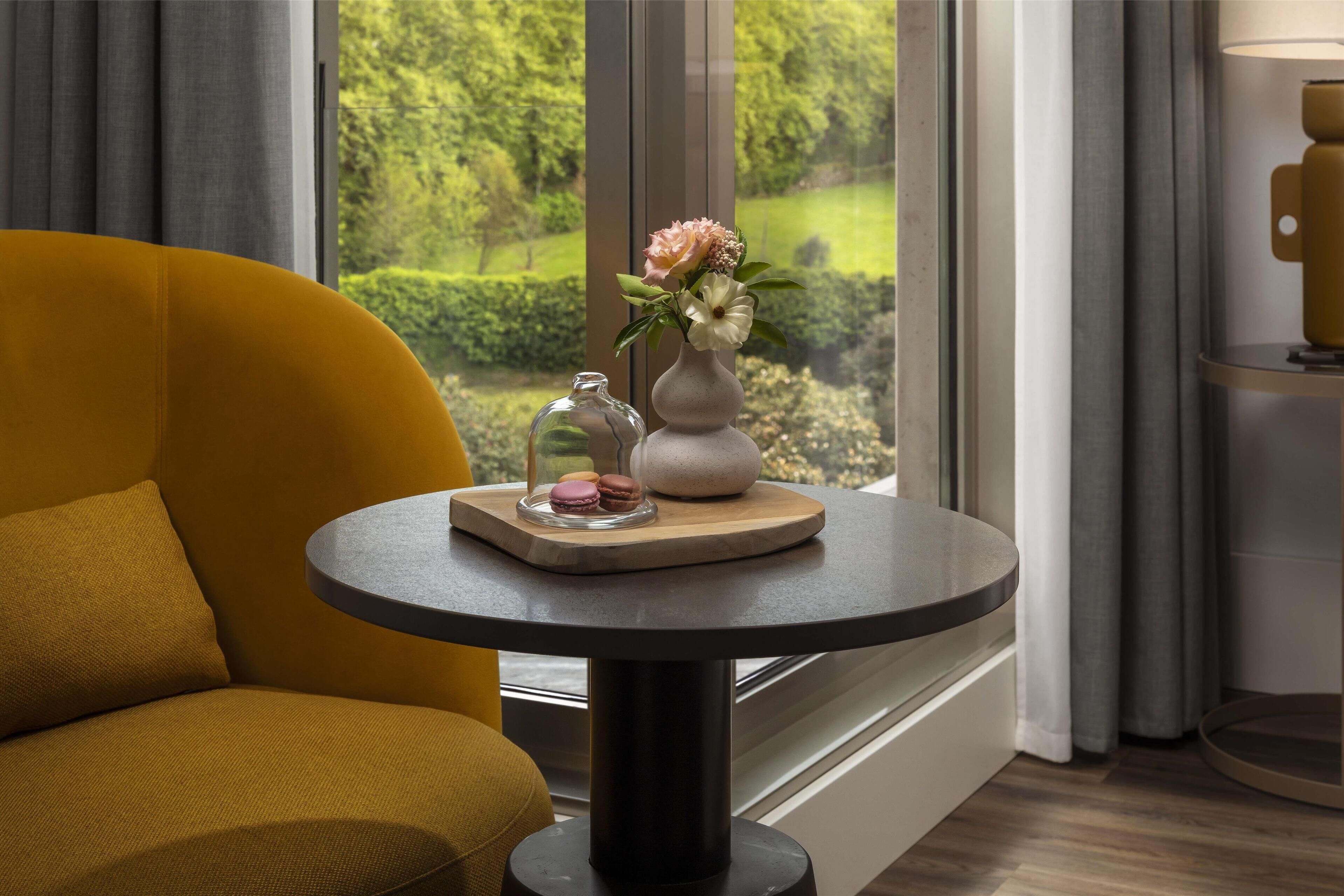 A yellow armchair, a round table with a vase and flowers, and a view of a garden through a window.