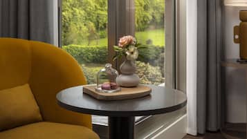 A yellow armchair, a round table with a vase and flowers, and a view of a garden through a window.