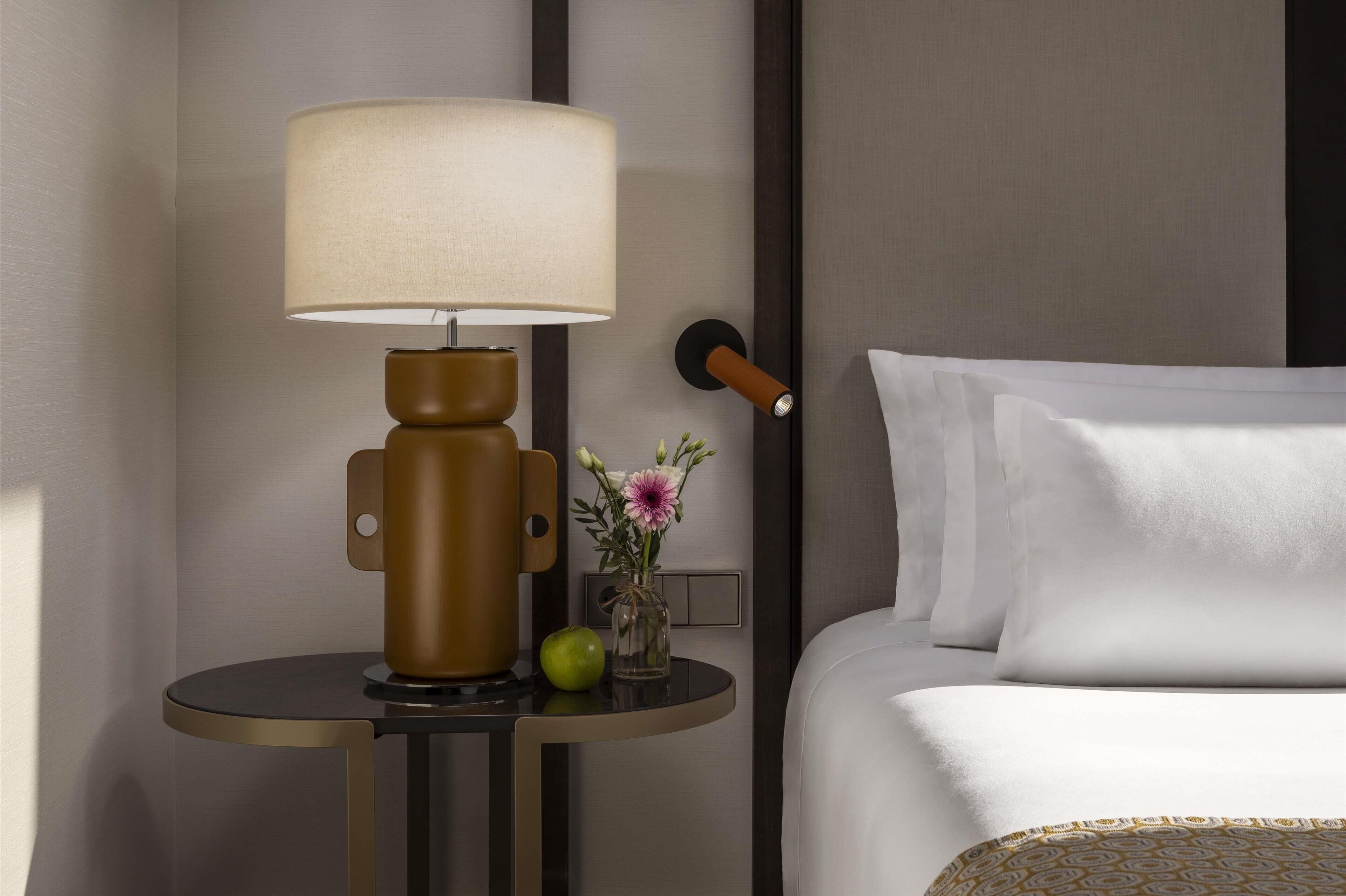 A hotel room with a bed, a round table with a lamp, a vase with flowers, and a green apple.