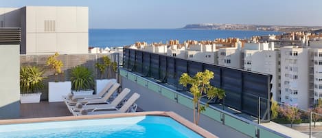 A rooftop pool with lounge chairs and potted plants, overlooking a cityscape and the sea.
