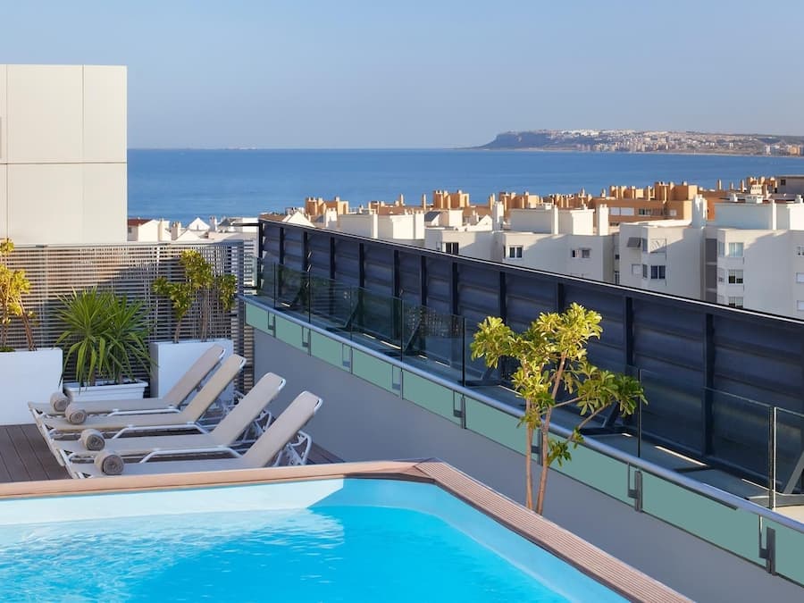 A rooftop pool with lounge chairs and potted plants, overlooking a cityscape and the sea.