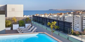 Una piscina en la azotea con sillones reclinables y plantas en macetas, con vistas a la ciudad y al mar.
