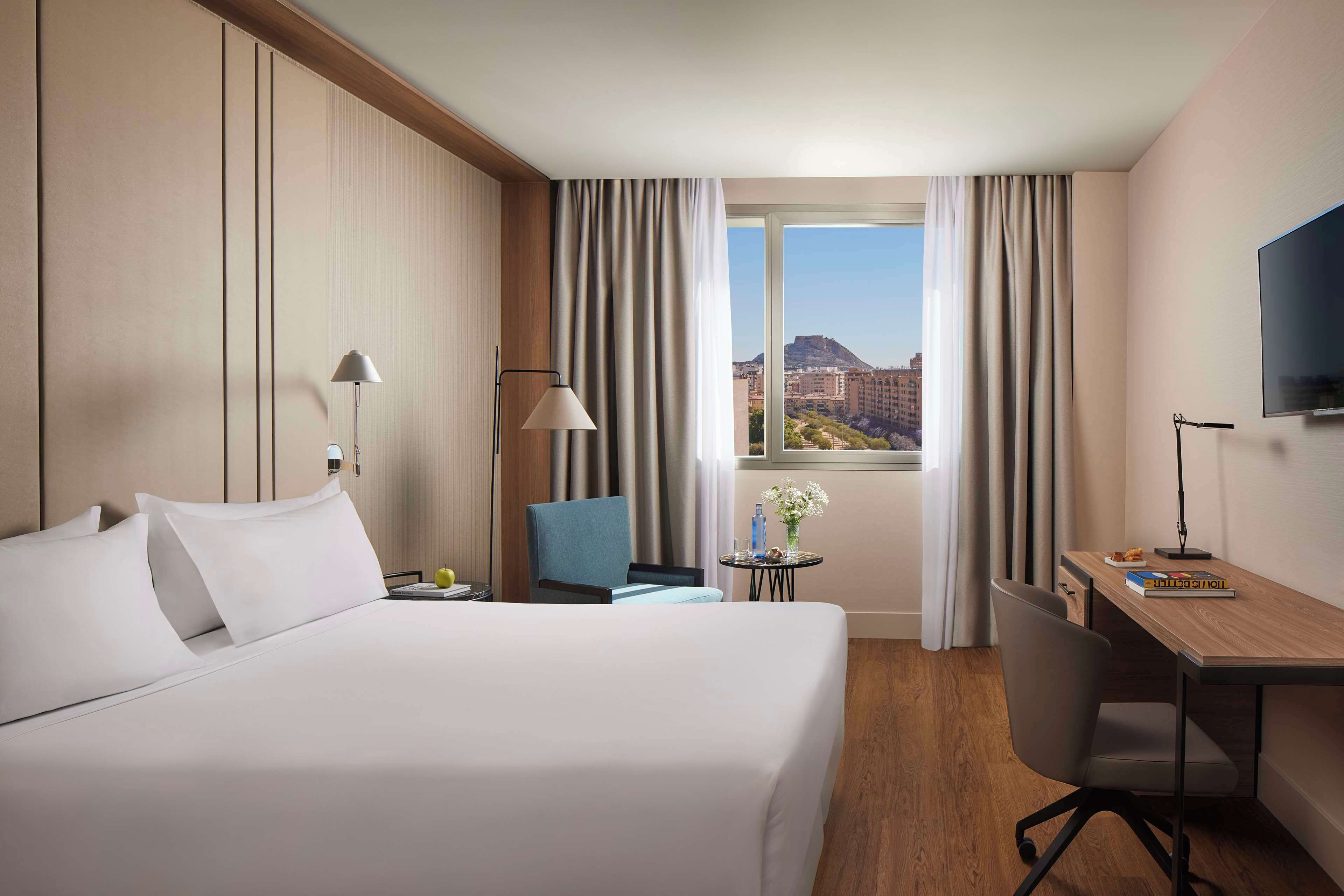 A hotel room with a large bed, a desk, a chair, a TV, and a window with curtains.
