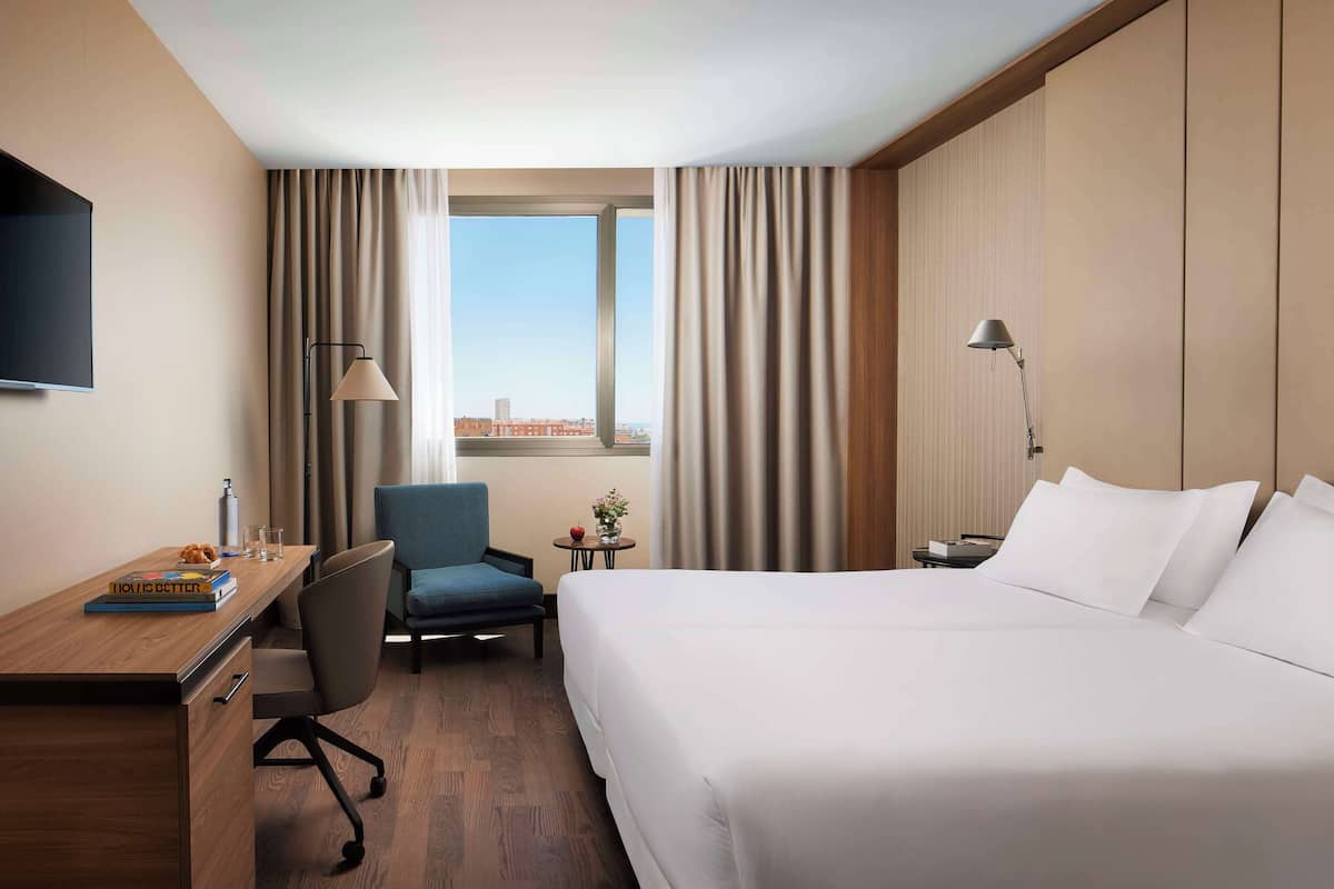 Ein Hotelzimmer mit Bett, Schreibtisch, Stuhl, Fernseher, Lampe und Blick auf die Stadt durch ein Fenster.