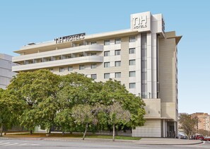 Un edificio hotelero de varios pisos con diseño moderno, que cuenta con balcones y una señalización llamativa de 'NH Hotels'.