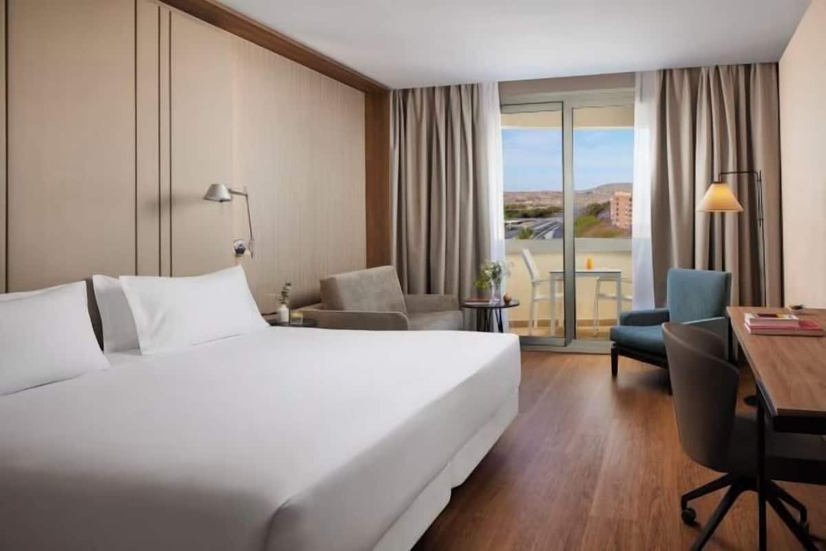 Ein Hotelzimmer mit einem großen Bett, einem Schreibtisch, einem Sessel und Blick auf die Stadtlandschaft.