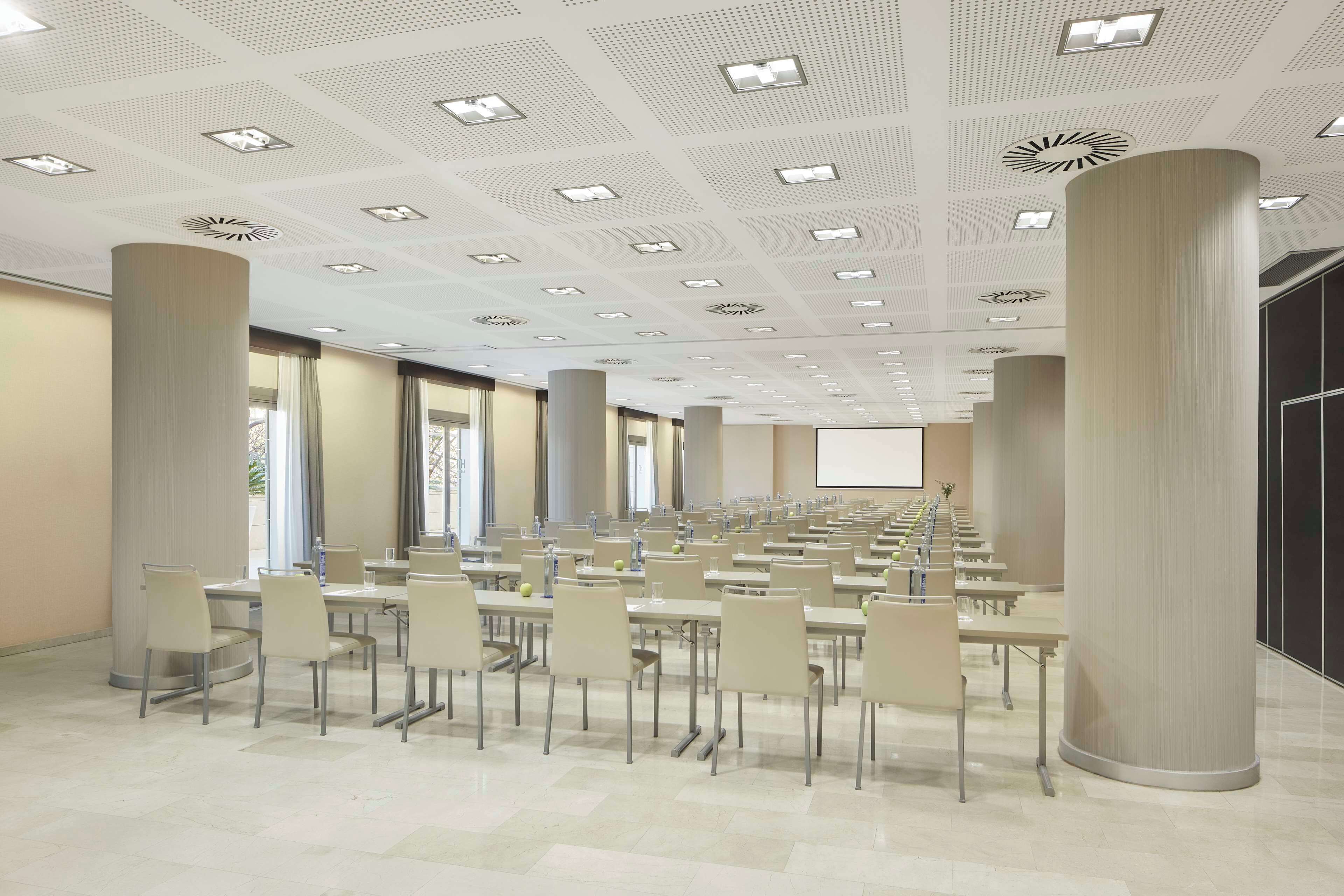 Een congreszaal met rijen tafels en stoelen, grote pilaren en een plafond met inbouwverlichting en ventilatie.
