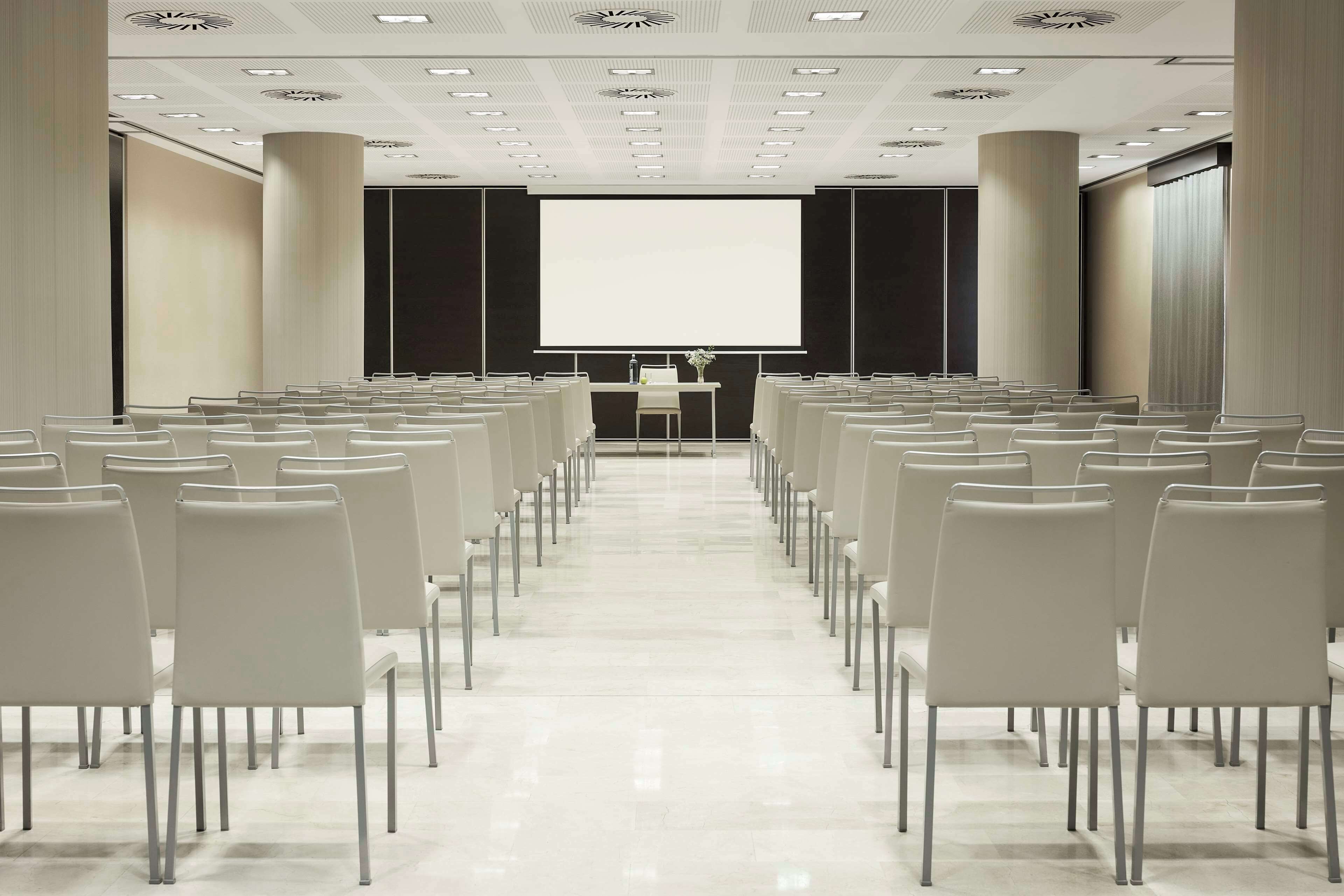 Une salle de conférence avec des rangées de chaises blanches, un grand écran à l’avant et un pupitre.