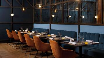Un restaurante con una larga mesa dispuesta para comer, asientos tapizados en azul y lámparas colgantes.