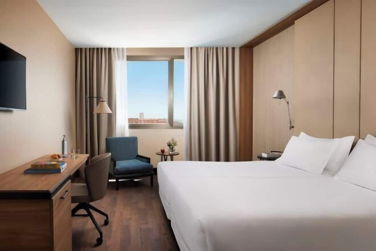 Ein Hotelzimmer mit Bett, Schreibtisch, Stuhl, Lampe und einem Fenster mit Vorhängen.