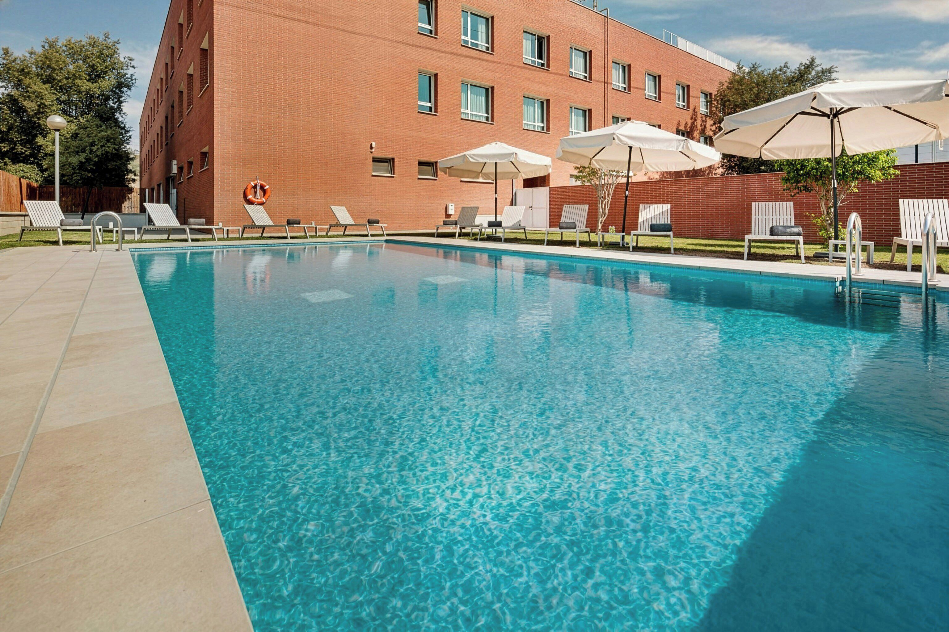 Piscina con sillas de descanso y sombrillas, rodeada por un edificio de ladrillo.