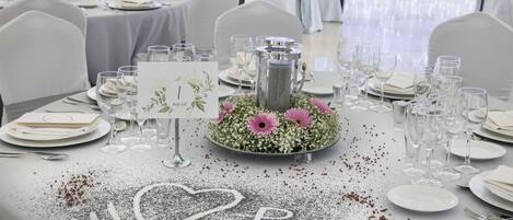 A wedding reception table set with white tablecloths, silverware, and wine glasses.