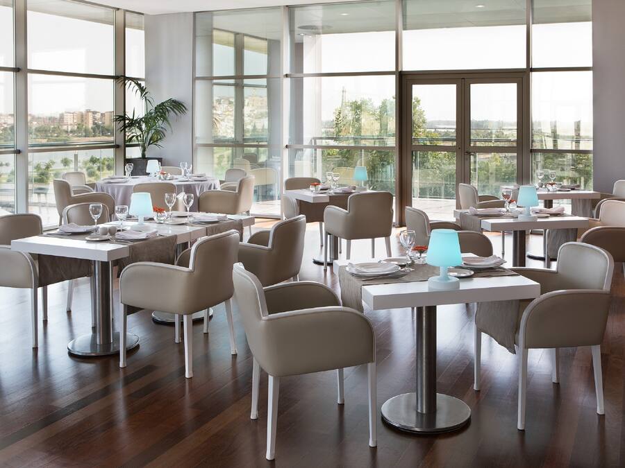 Un comedor moderno con varias mesas y sillas, ventanas amplias que ofrecen vistas del paisaje urbano, y una planta en maceta en la esquina.