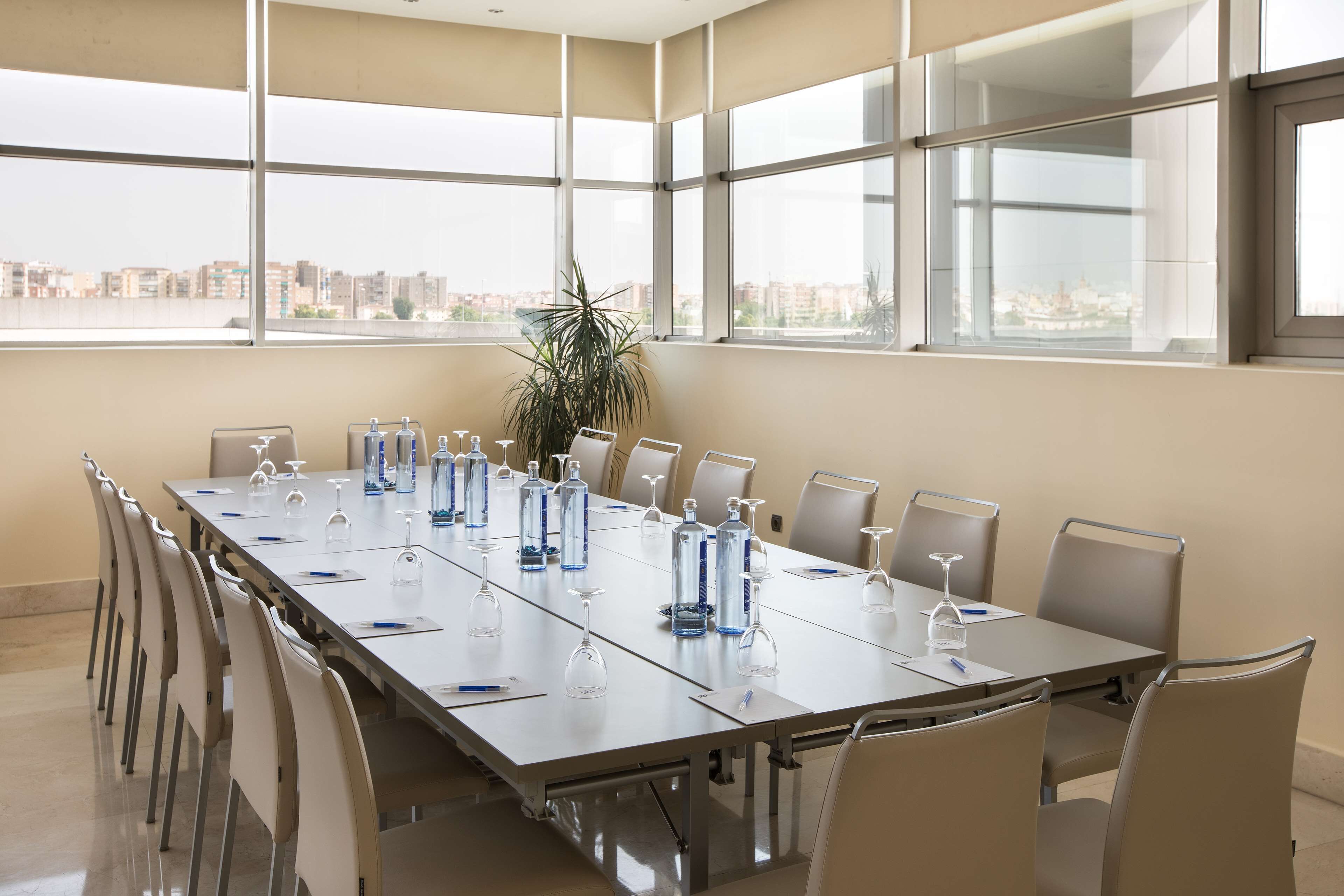 Un salón de conferencias con una mesa larga, sillas, botellas de agua, copas de vino y una planta en maceta.