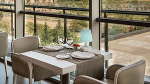 Mesa de comedor lista con platos, vasos y una lámpara, con vista a un paisaje urbano que incluye un río y un puente.