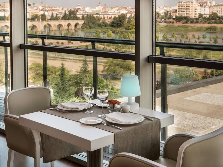 Mesa de comedor lista con platos, vasos y una lámpara, con vista a un paisaje urbano que incluye un río y un puente.