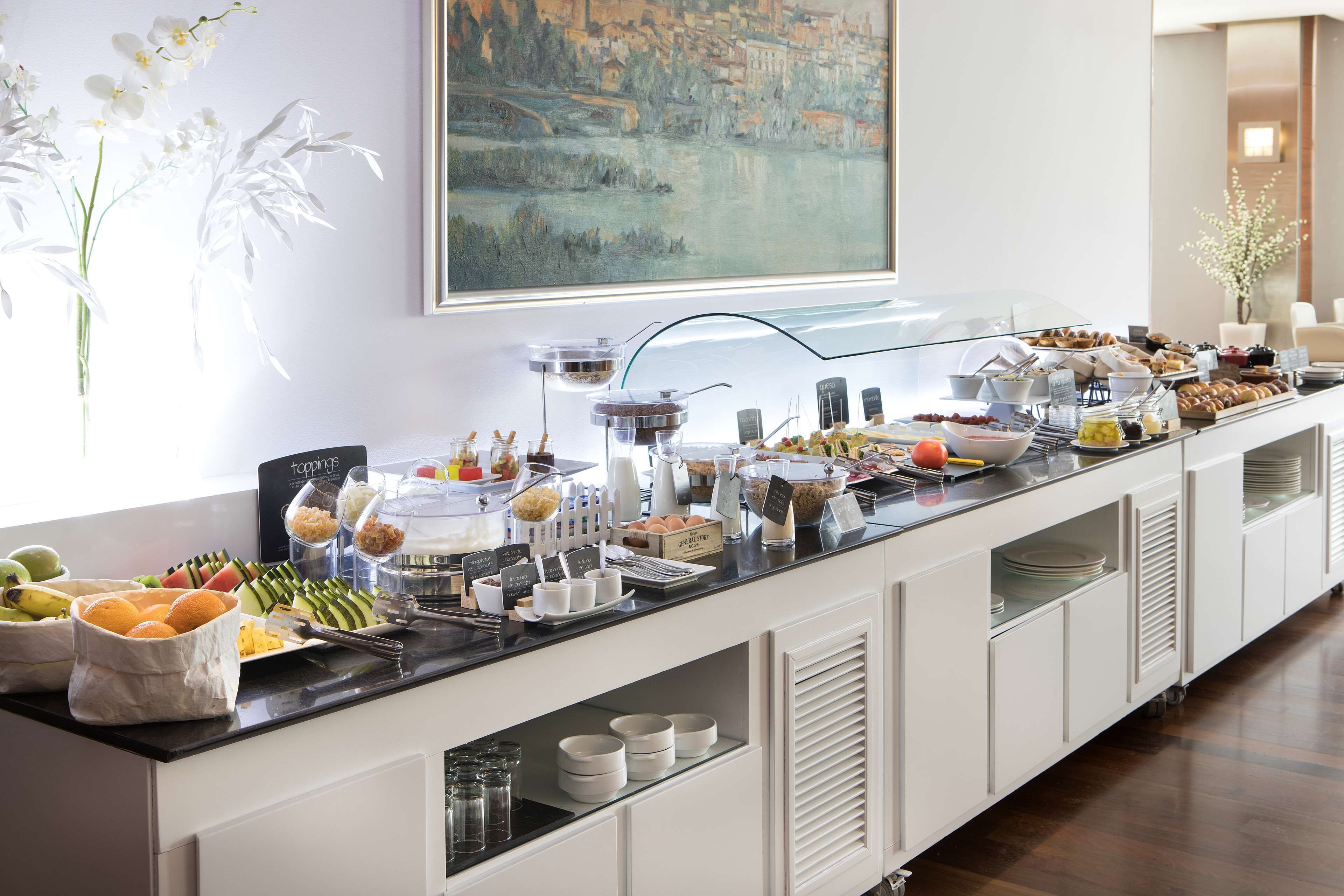 A buffet setup with various food items and condiments, a painting on the wall, and a vase with white flowers.