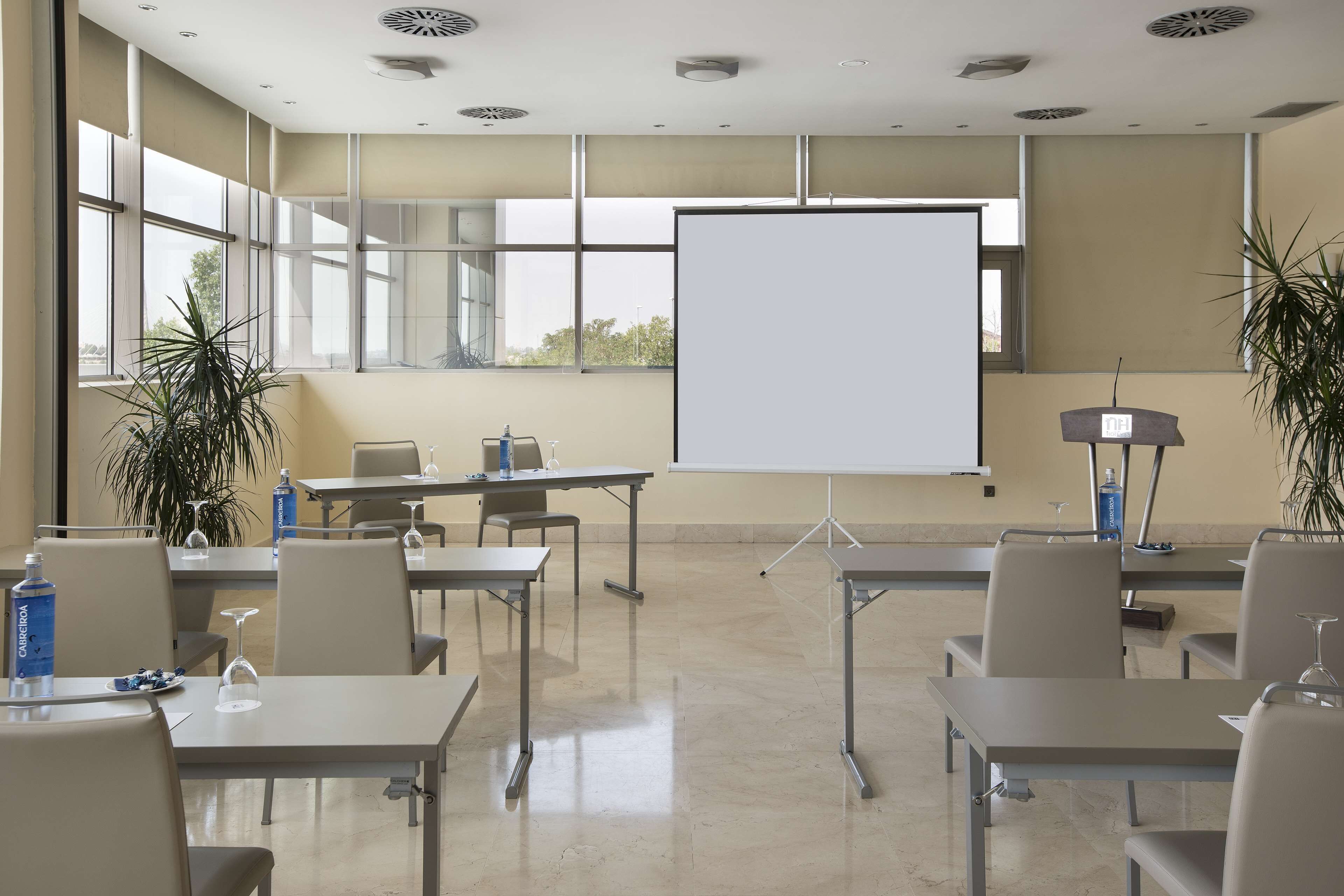 Un salón de conferencias con mesas, sillas, una pantalla de proyector y plantas en macetas.
