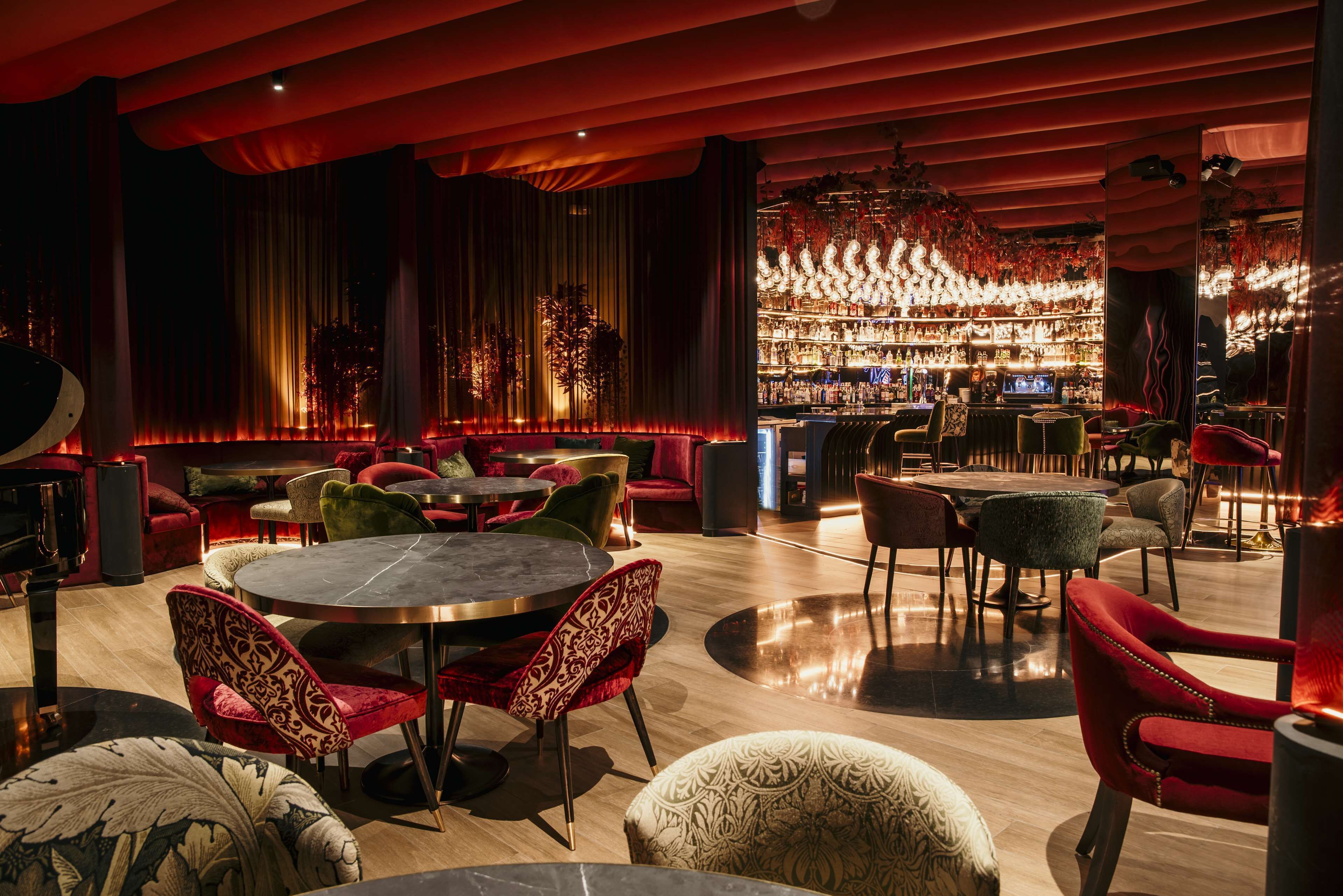 A bar area with round tables and red chairs, a bar counter with a variety of bottles, and a large chandelier.