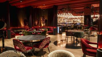 A bar area with round tables and red chairs, a bar counter with a variety of bottles, and a large chandelier.