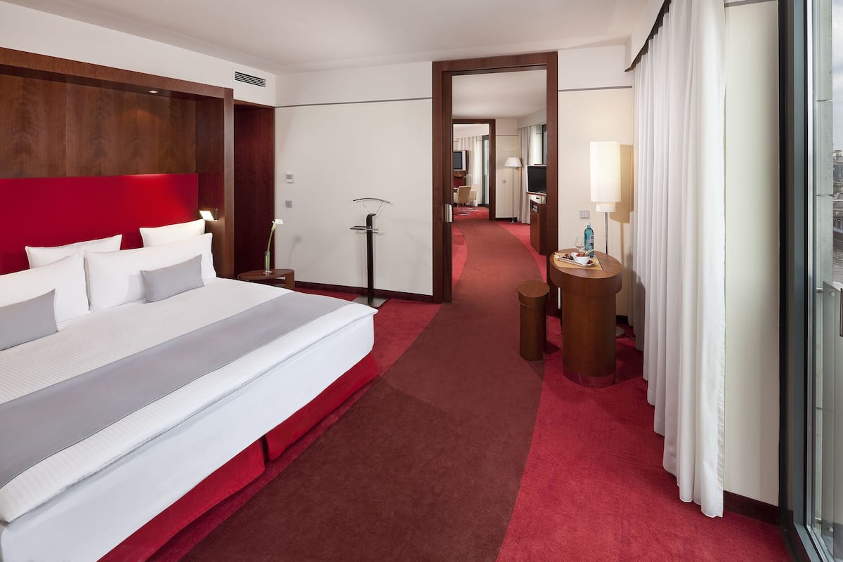 A hotel room with a large bed, a desk with a lamp, a TV, and a view of the city through the window.
