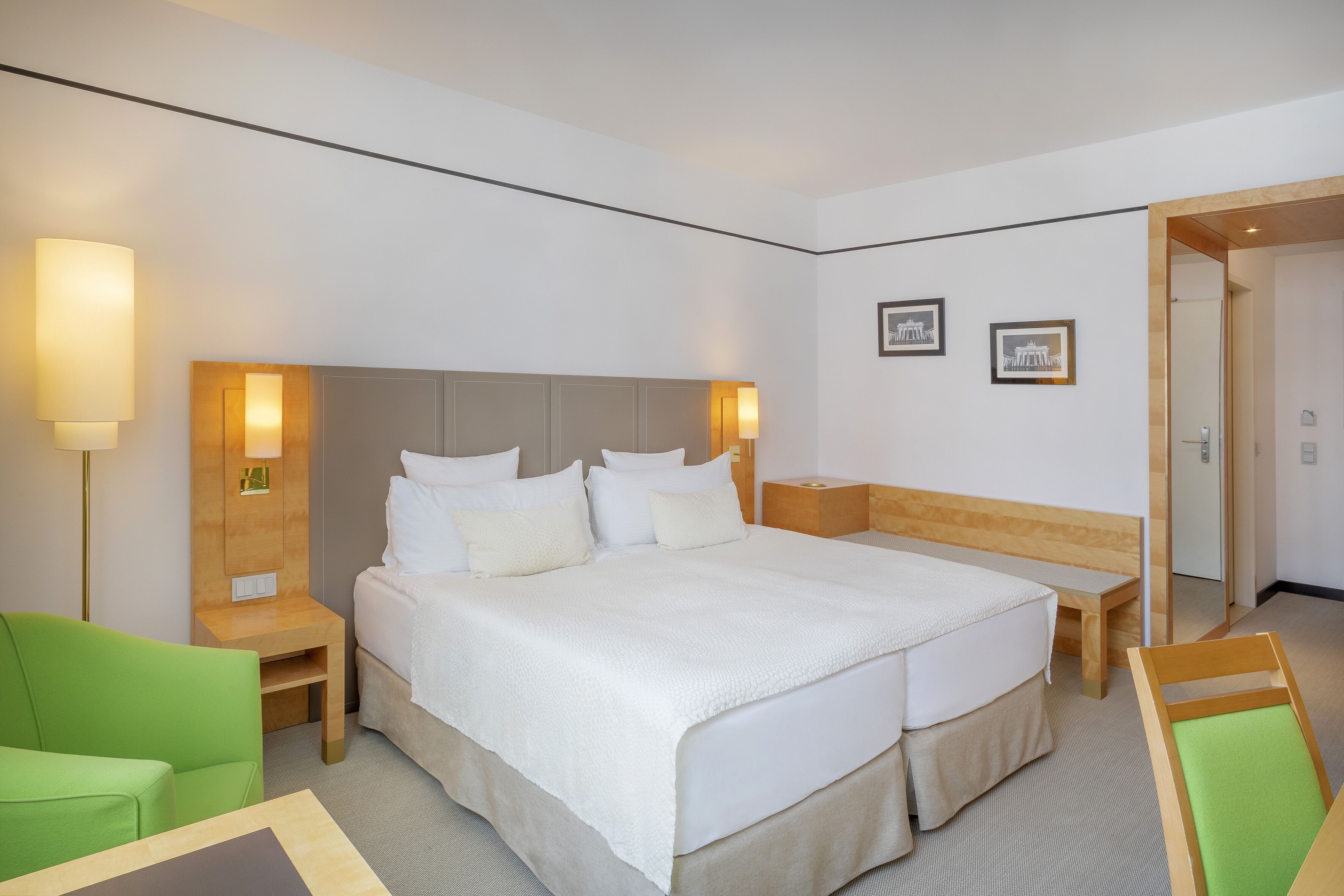 A hotel room with a large bed, bedside tables, a green armchair, and a wooden bench.