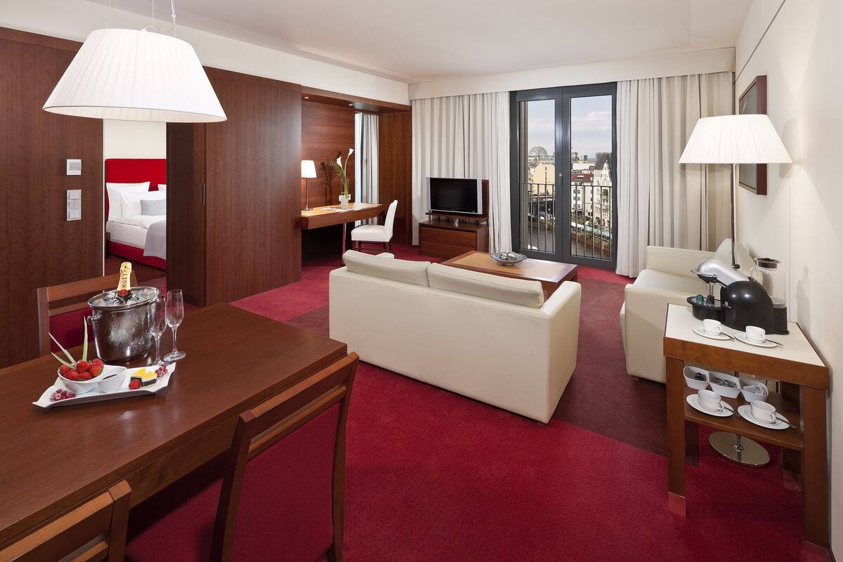 A hotel room with a dining area, a living room, a TV, a balcony with a city view, and a minibar.