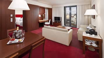 A hotel room with a dining area, a living room, a TV, a balcony with a city view, and a minibar.