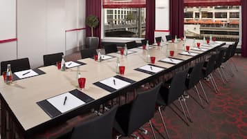 Un salón de conferencias con una larga mesa, sillas negras, cortinas rojas y blancas, y vistas a un puente y edificios a través de amplias ventanas.