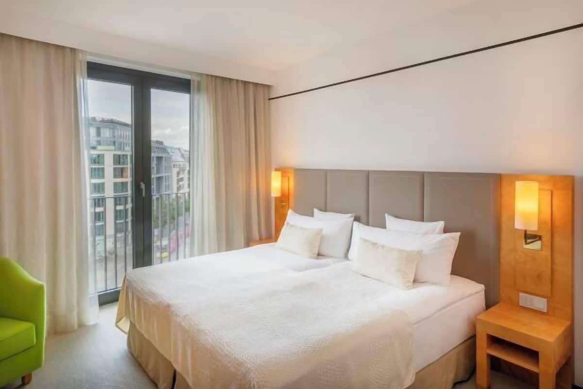 A hotel room with a large bed, two bedside tables, a green chair, and a view of buildings through the window.