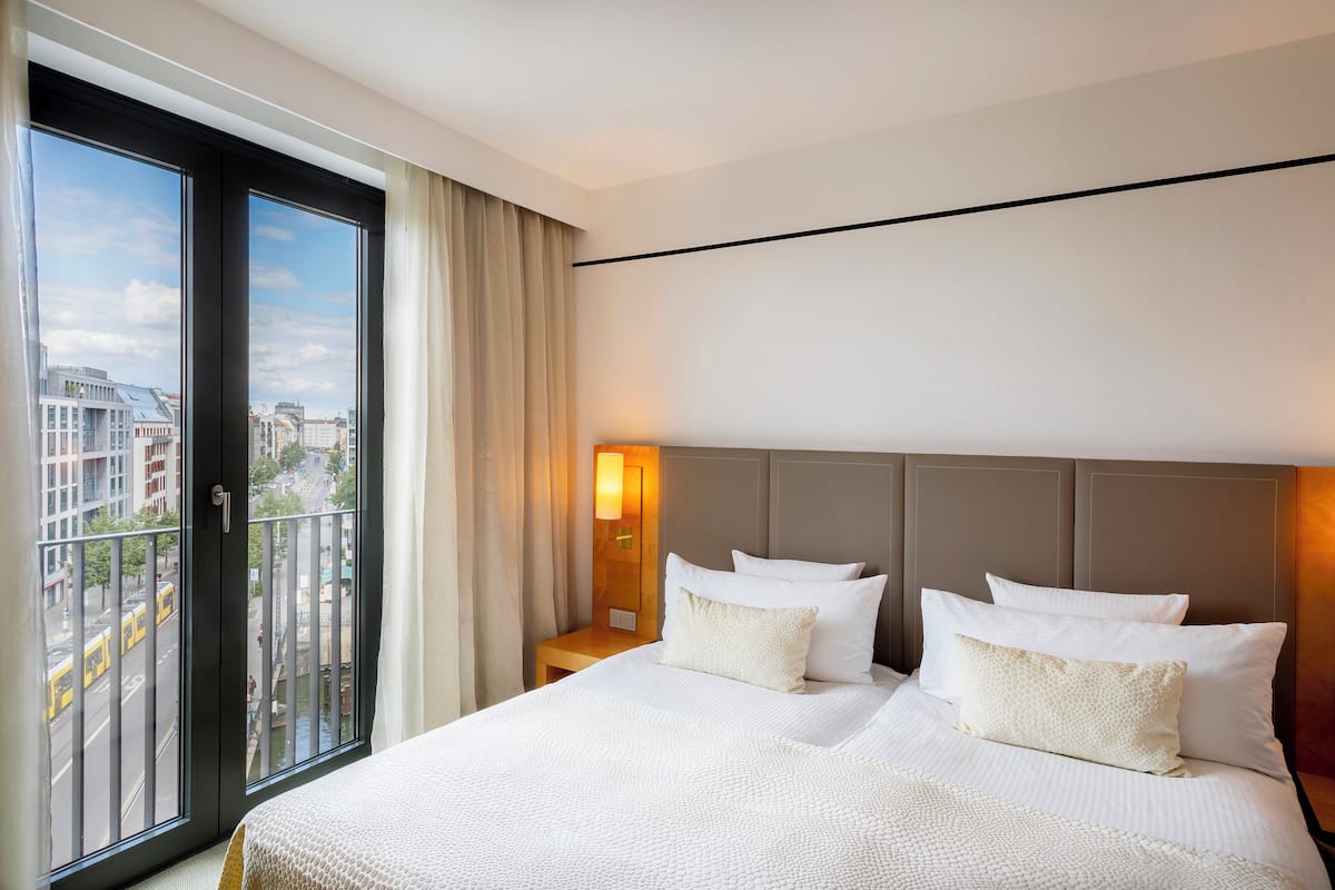 A hotel room with a large bed, a nightstand with a lamp, and a view of the city through a large window.