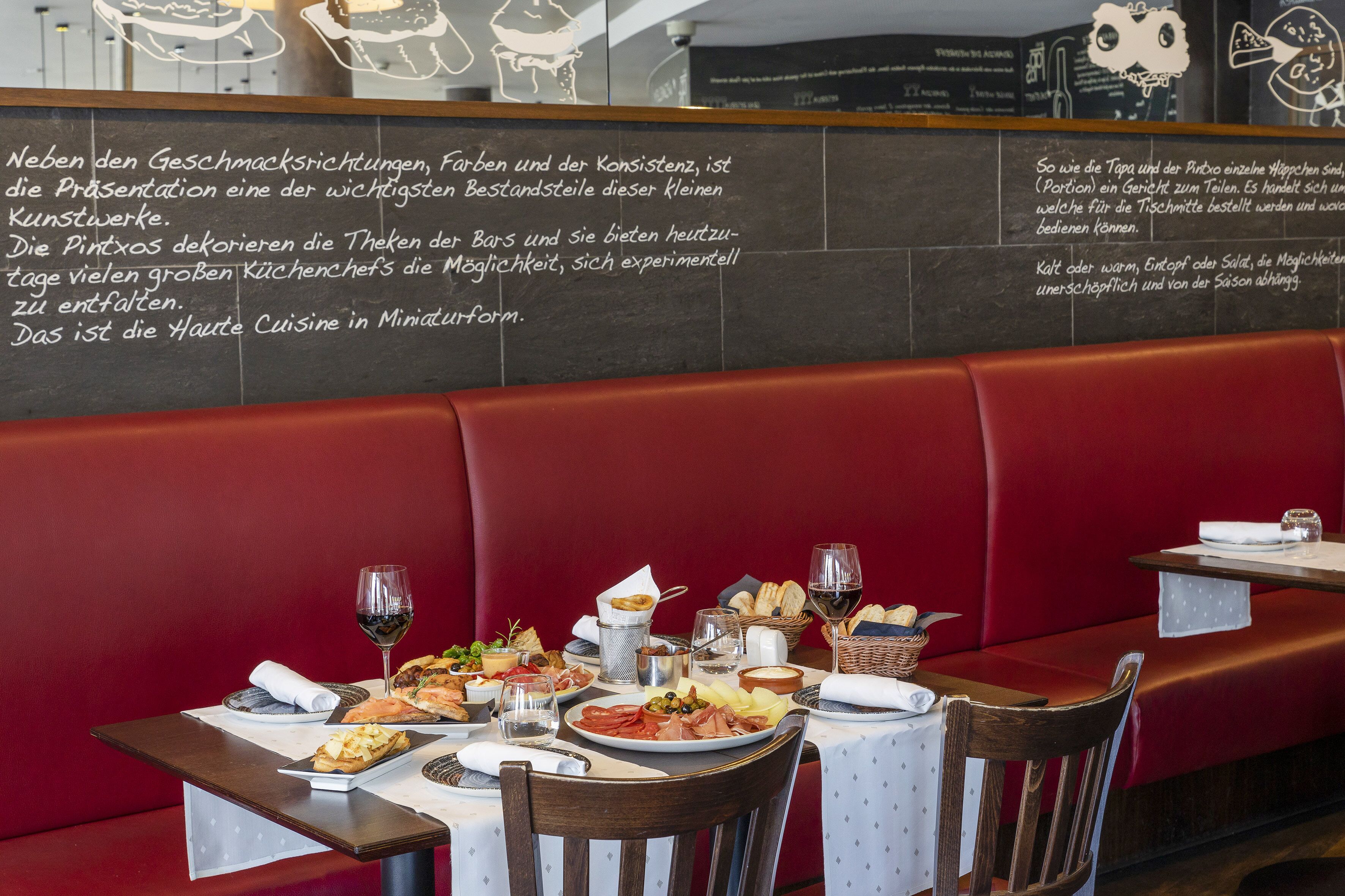 A restaurant interior with a red booth seating area, a table set with various dishes, wine glasses, and a chalkboard menu.