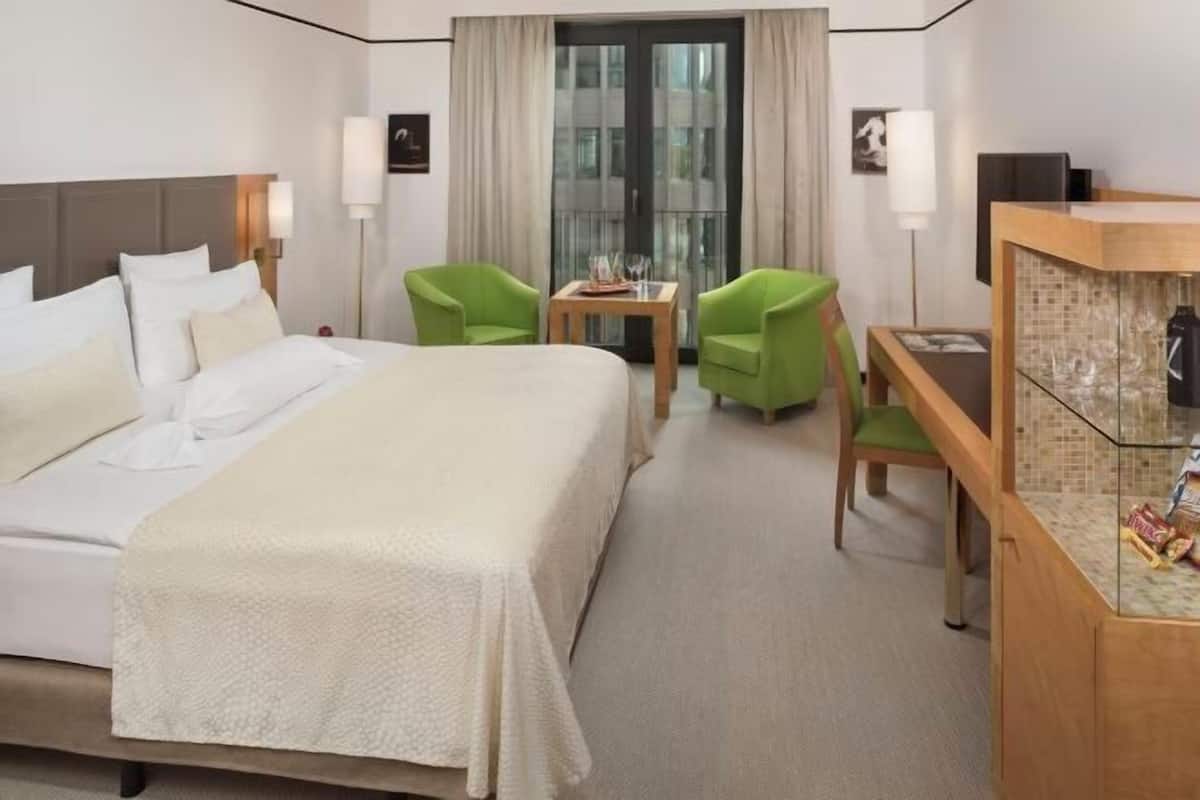 A hotel room with a bed, two green chairs, a small table, a minibar, and a view through a glass door.