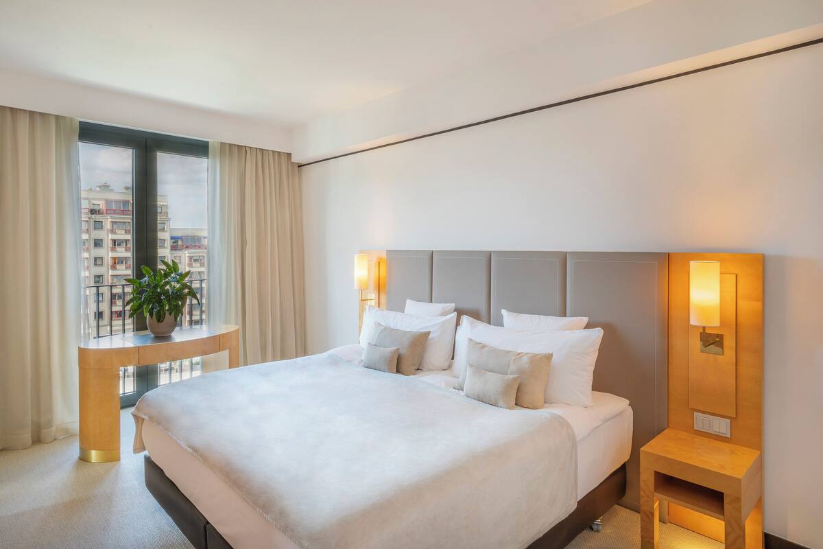 A hotel room with a large bed, bedside lamps, a small table, and a view of buildings through the window.