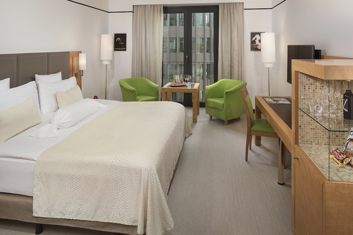 A hotel room with a large bed, two green chairs, a small table with wine glasses, a minibar, and a large window with curtains.