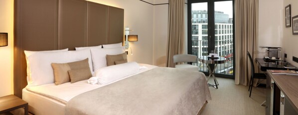 A hotel room with a large bed, a desk, a chair, and a view of the city through the window.