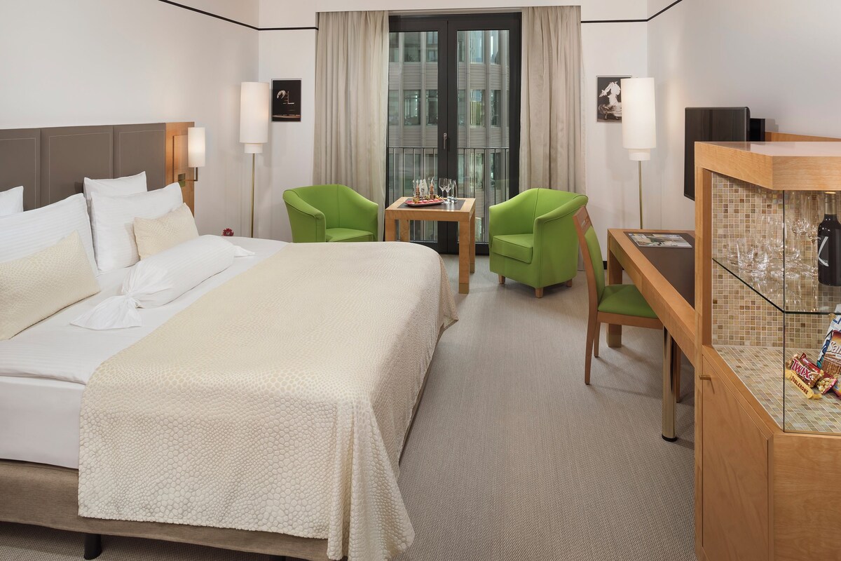 A hotel room with a large bed, two green chairs, a small table with wine glasses, a minibar, and a large window with curtains.