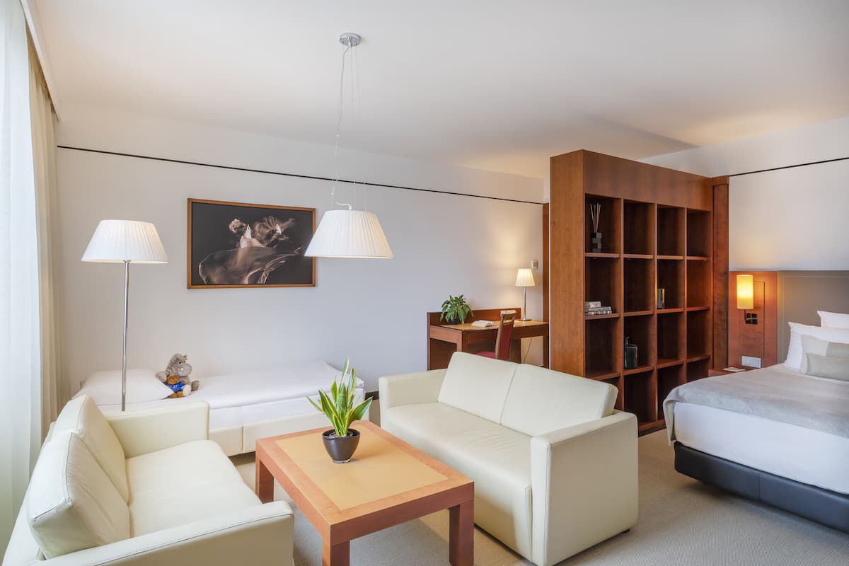 A hotel room with a bed, two white sofas, a wooden coffee table, a desk, a bookshelf, and a potted plant.