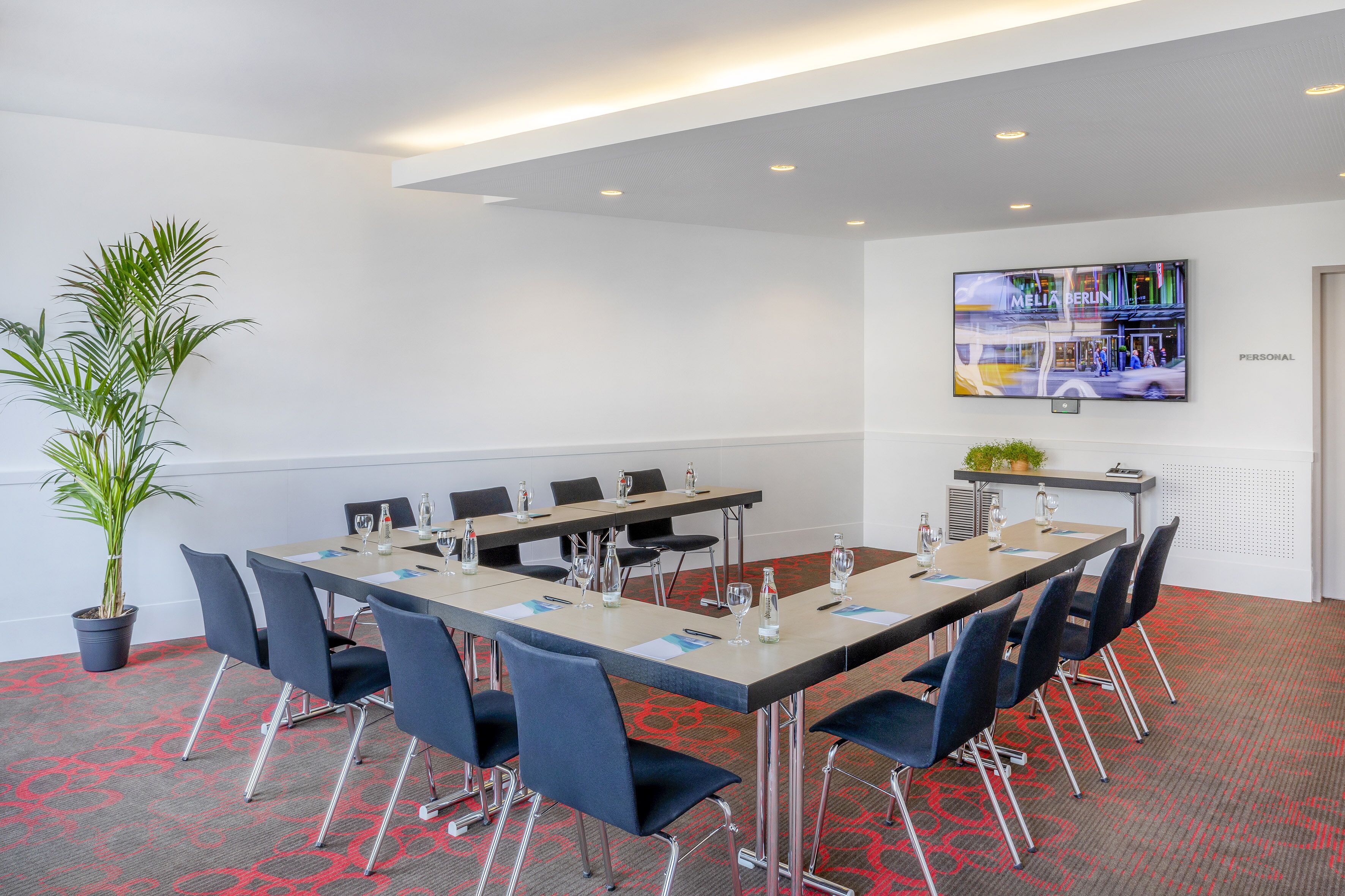 A conference room with a long table, chairs, a potted plant, and a TV screen displaying 'MELIA BERLIN'.